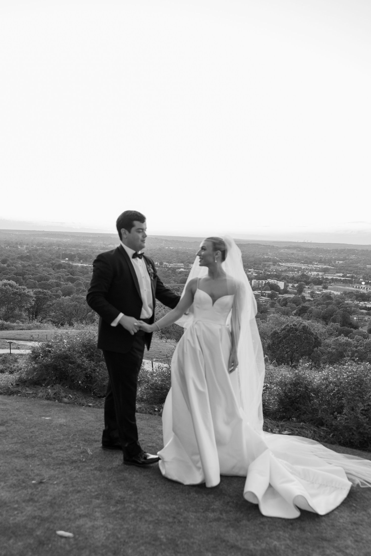 A bride and groom share a romantic moment on a hilltop overlooking a scenic landscape in black and white photography.