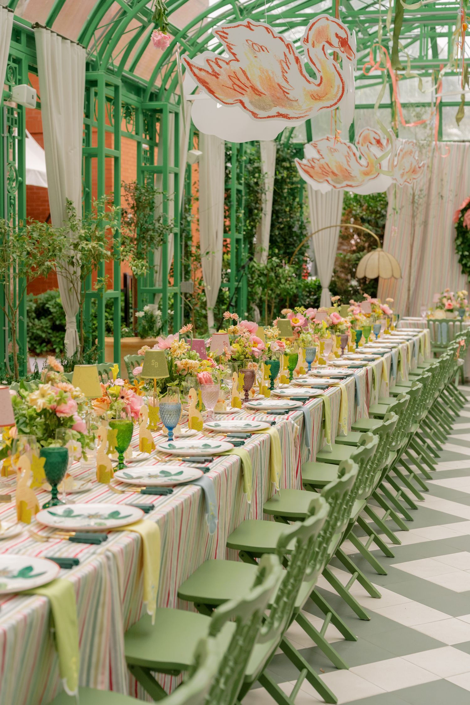 Elegant greenhouse dining with green chairs, striped tablecloth, colorful florals, and swan decorations overhead.