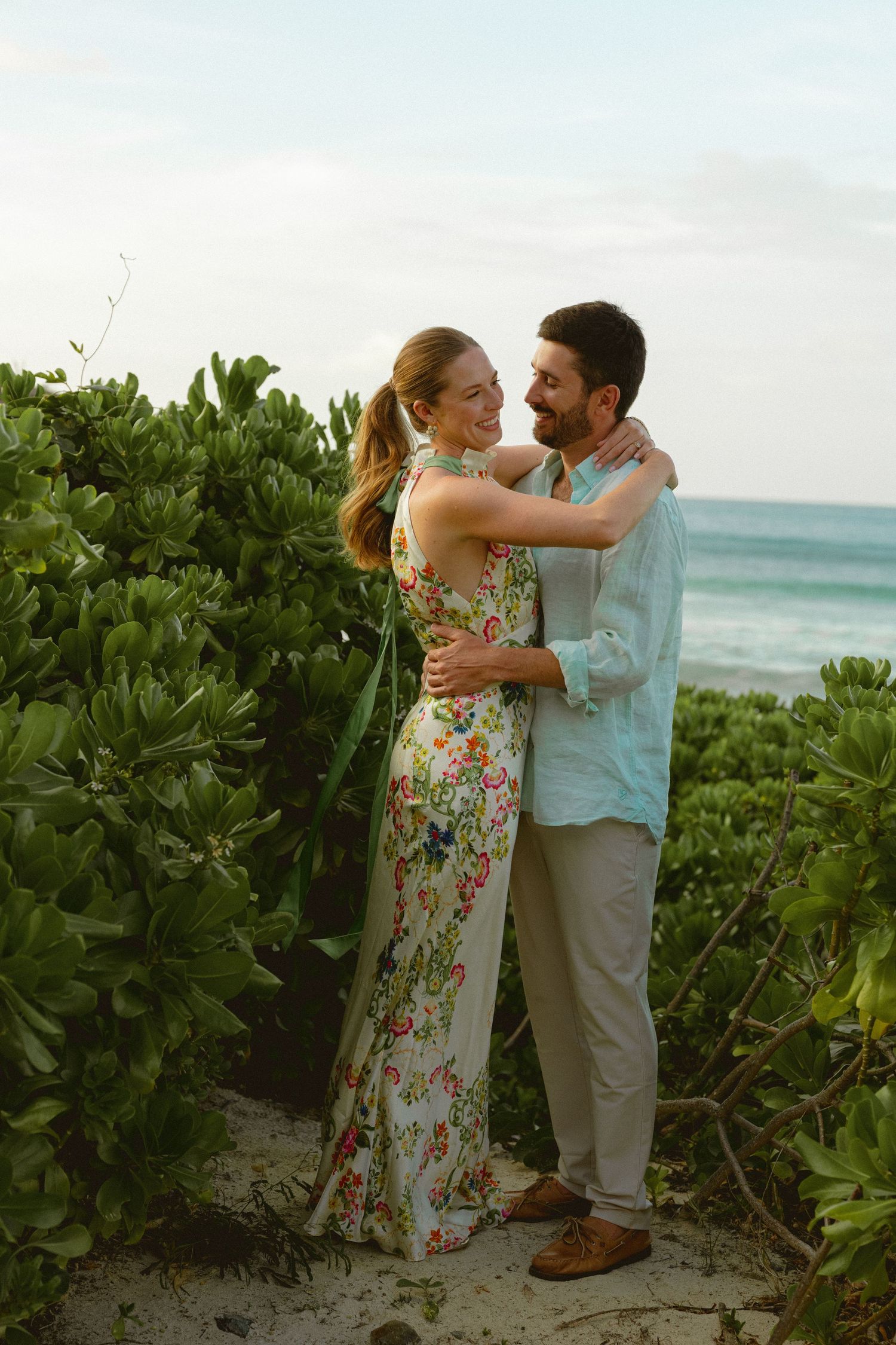 Couple embracing near lush greenery with ocean backdrop, woman in floral dress, man in light blue shirt.