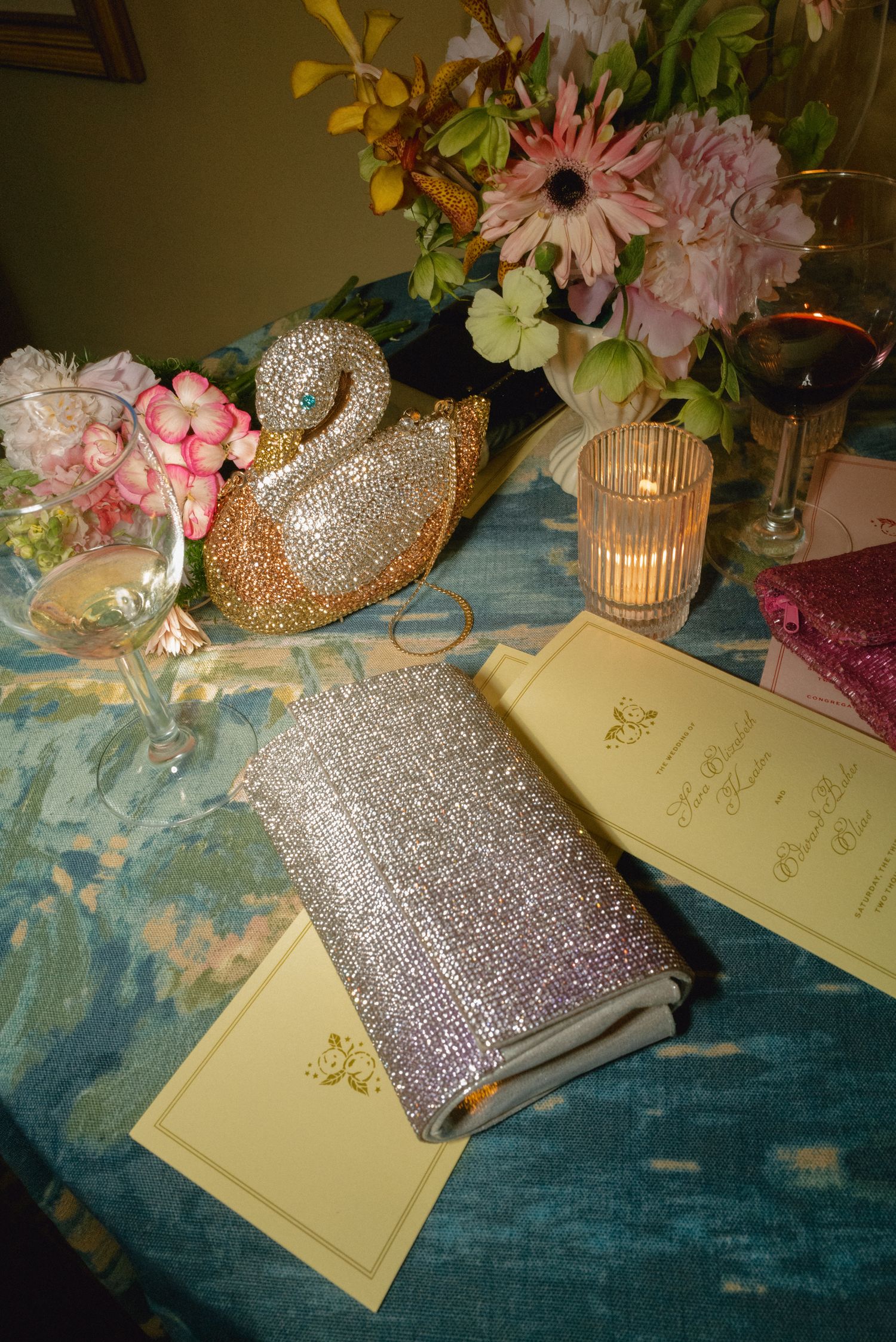 Elegant table setting with crystal swan clutch, sparkly purse, floral arrangement, candle, and wine glass on blue tablecloth.