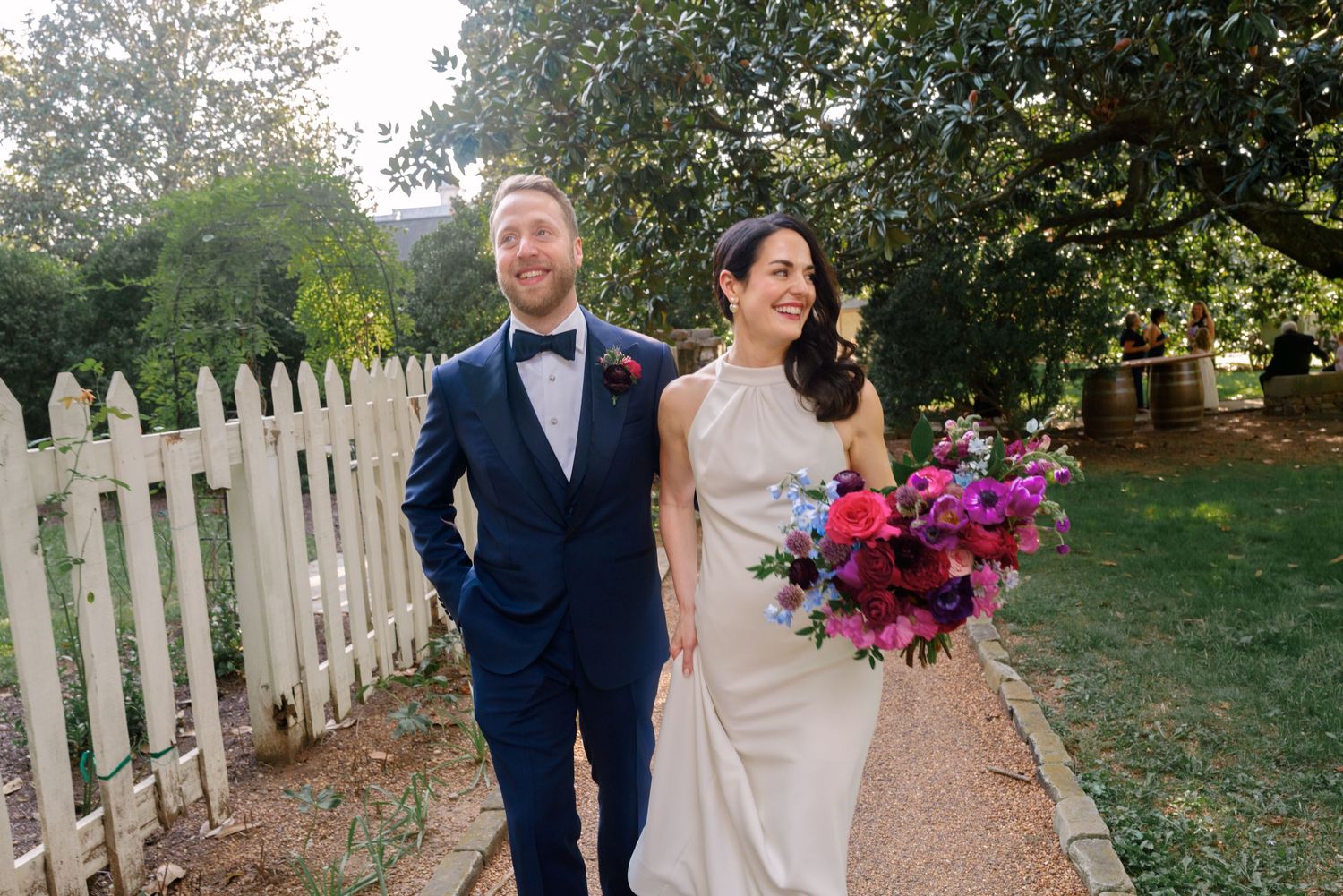 Newlyweds walk along garden path with white picket fence as bride holds vibrant purple and pink bouquet.