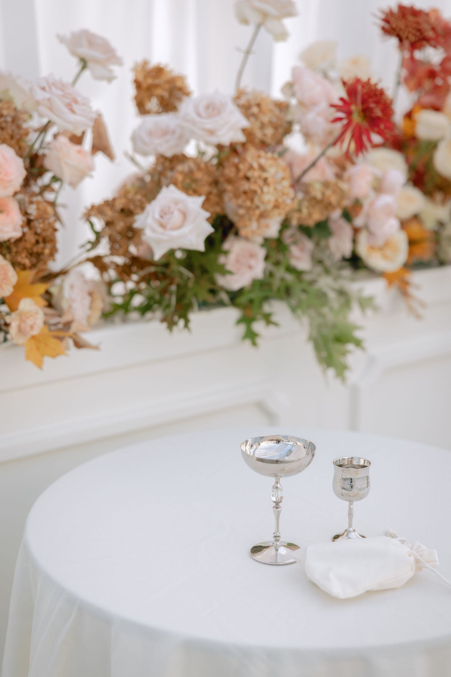 Silver chalices and a ring dish on a white draped table with a lush floral arrangement of roses and hydrangeas.