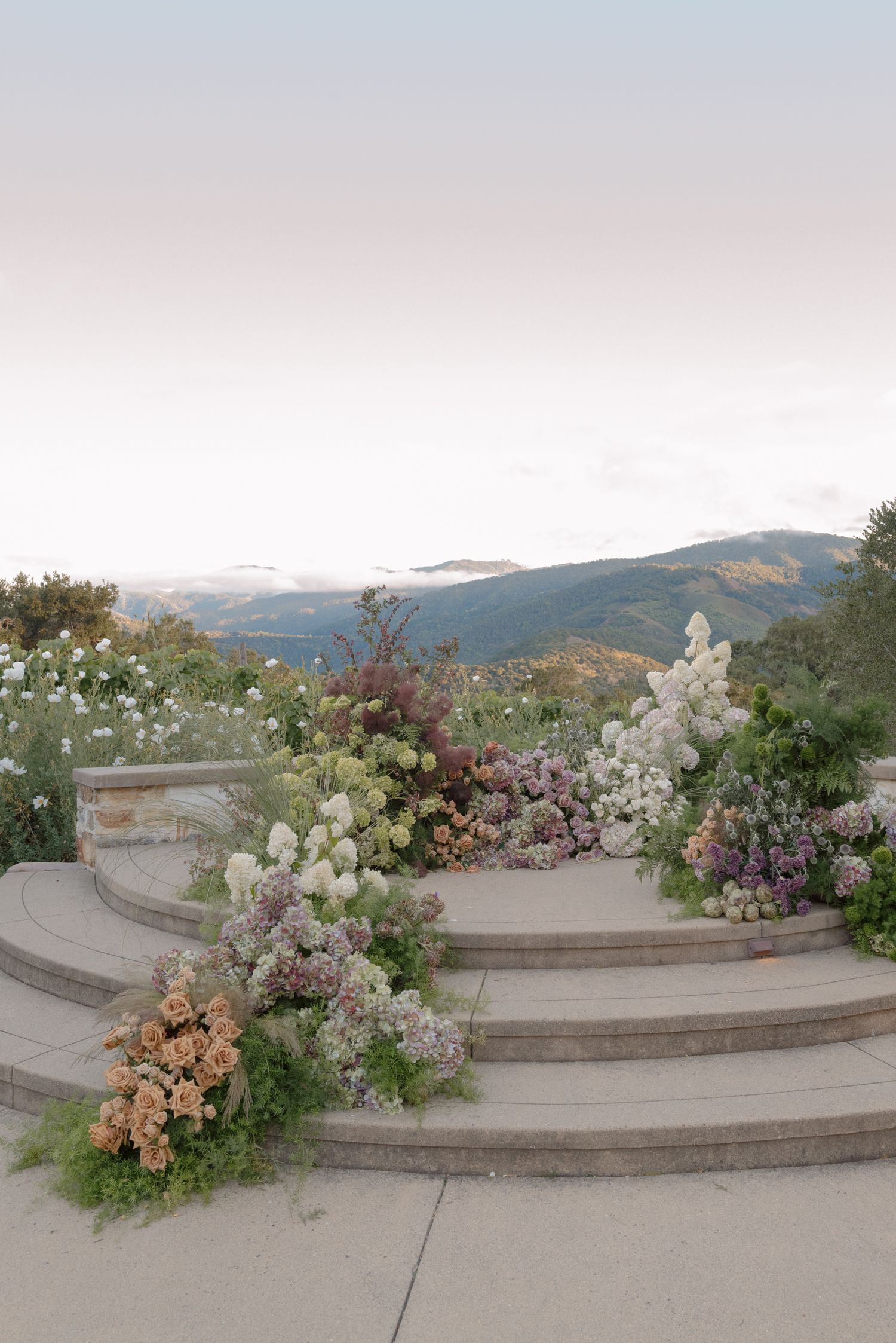 Lush floral arrangements in soft pastels cascade down stone steps with misty mountain views in the background.