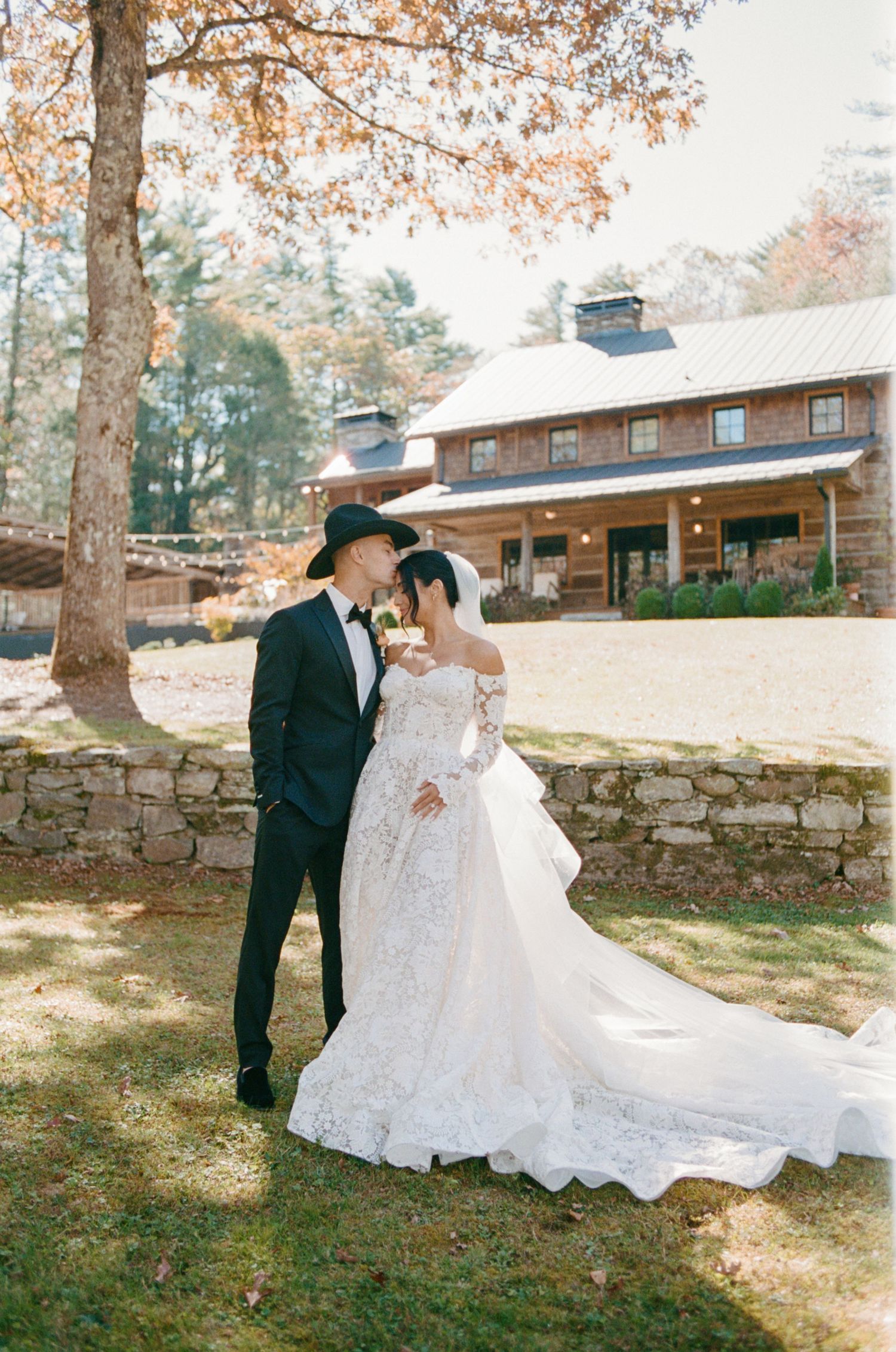 Bride and groom in cowboy hat share romantic moment at rustic barn wedding venue surrounded by autumn foliage.