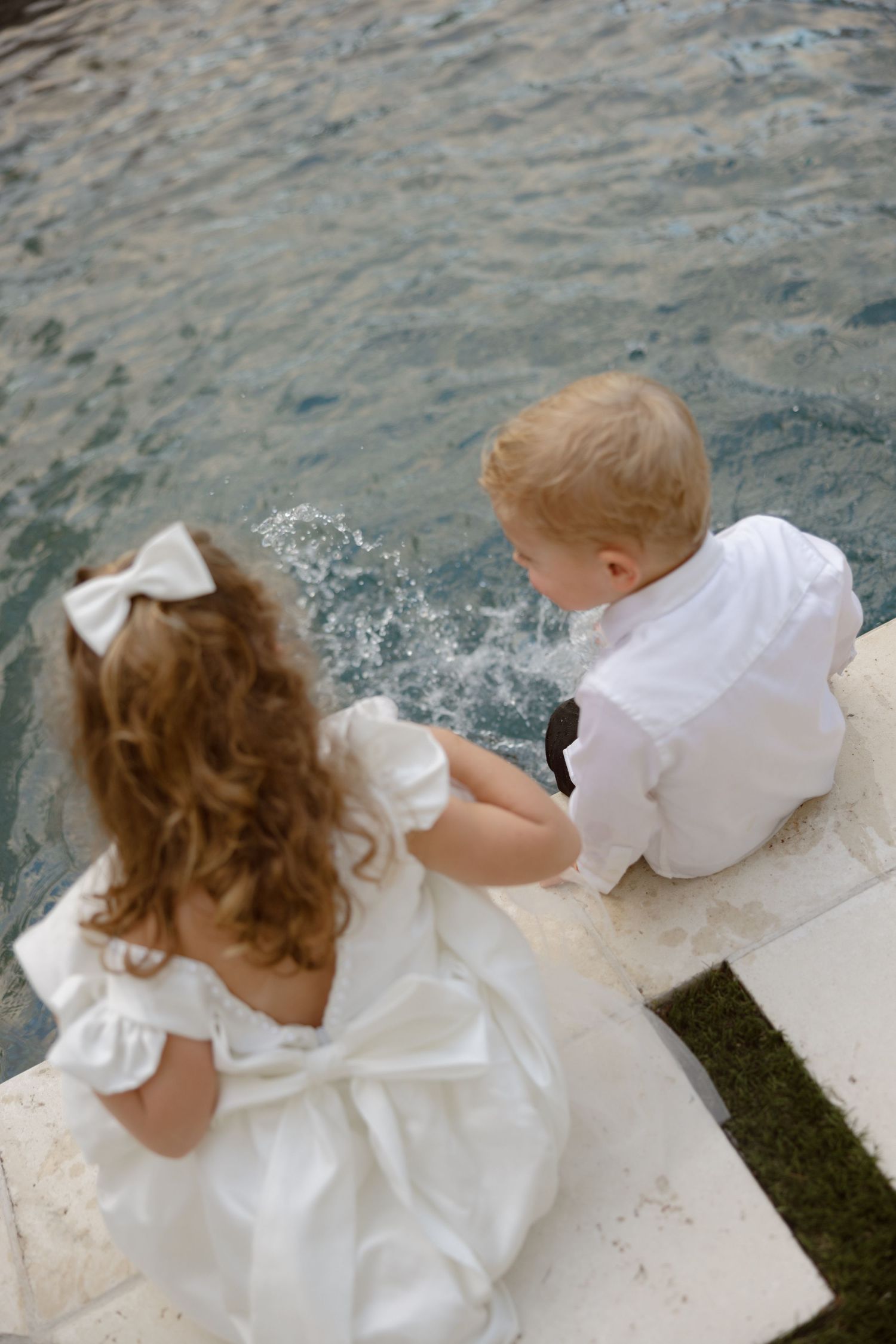 Two young children in white formal attire sitting by a fountain, viewed from above.
