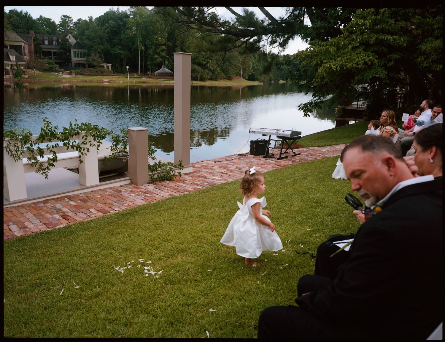A flower girl in a white dress walks on green grass near a lakeside wedding ceremony with seated guests.