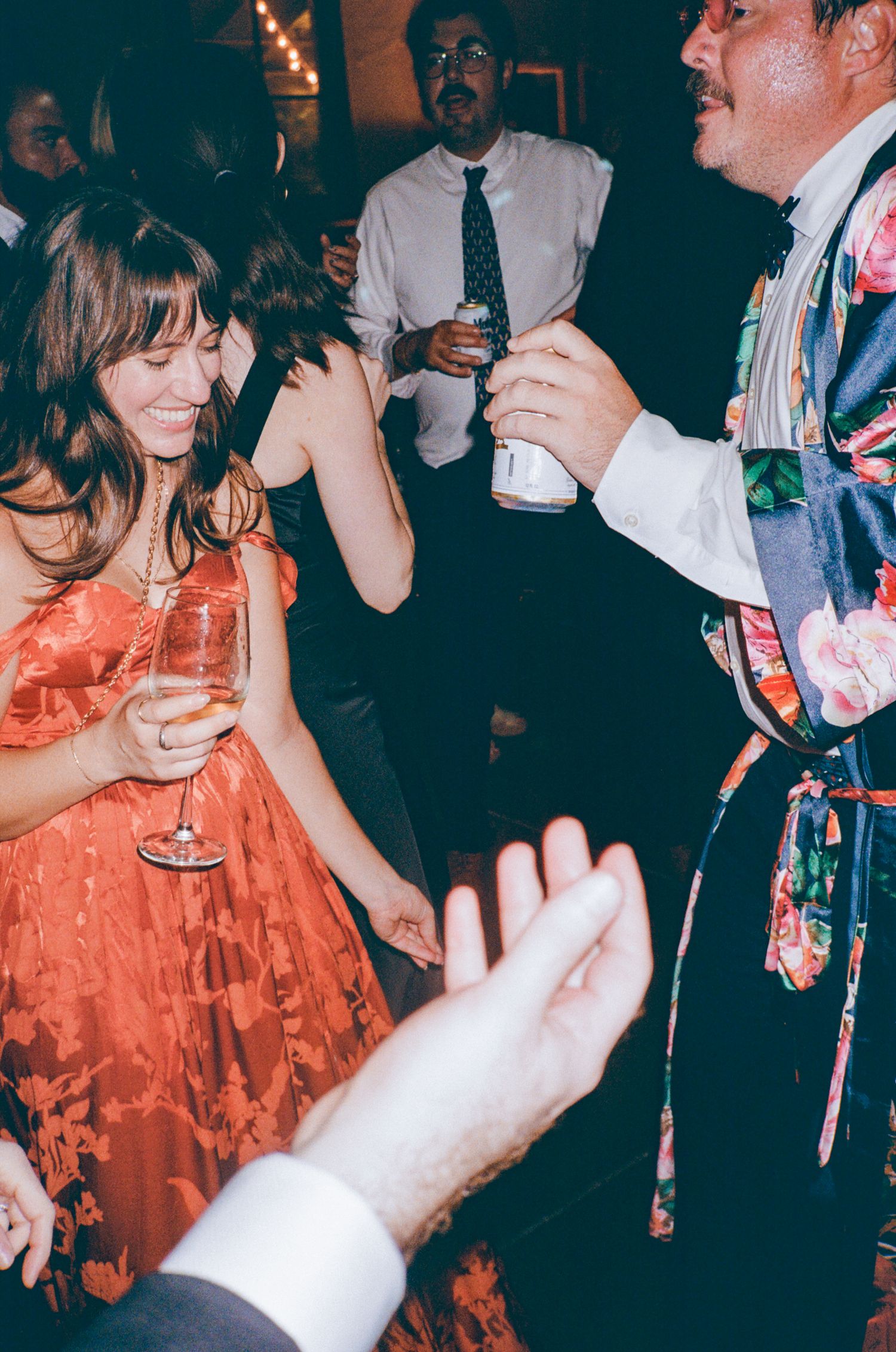 Partygoers in formal attire dance and socialize while holding drinks at a dimly lit evening celebration.