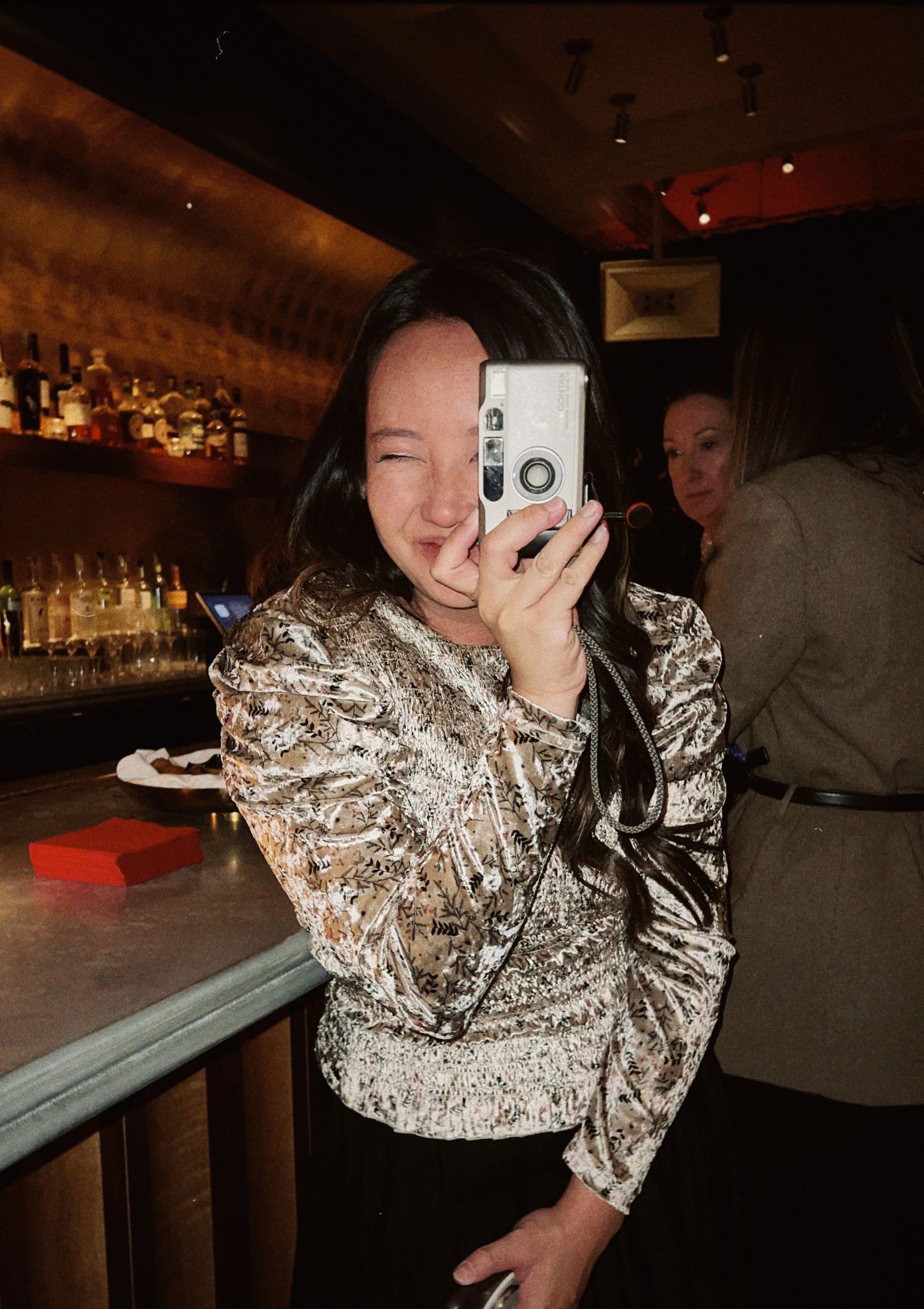 A young woman in a sequined top takes a selfie with a film camera at a dimly lit bar.