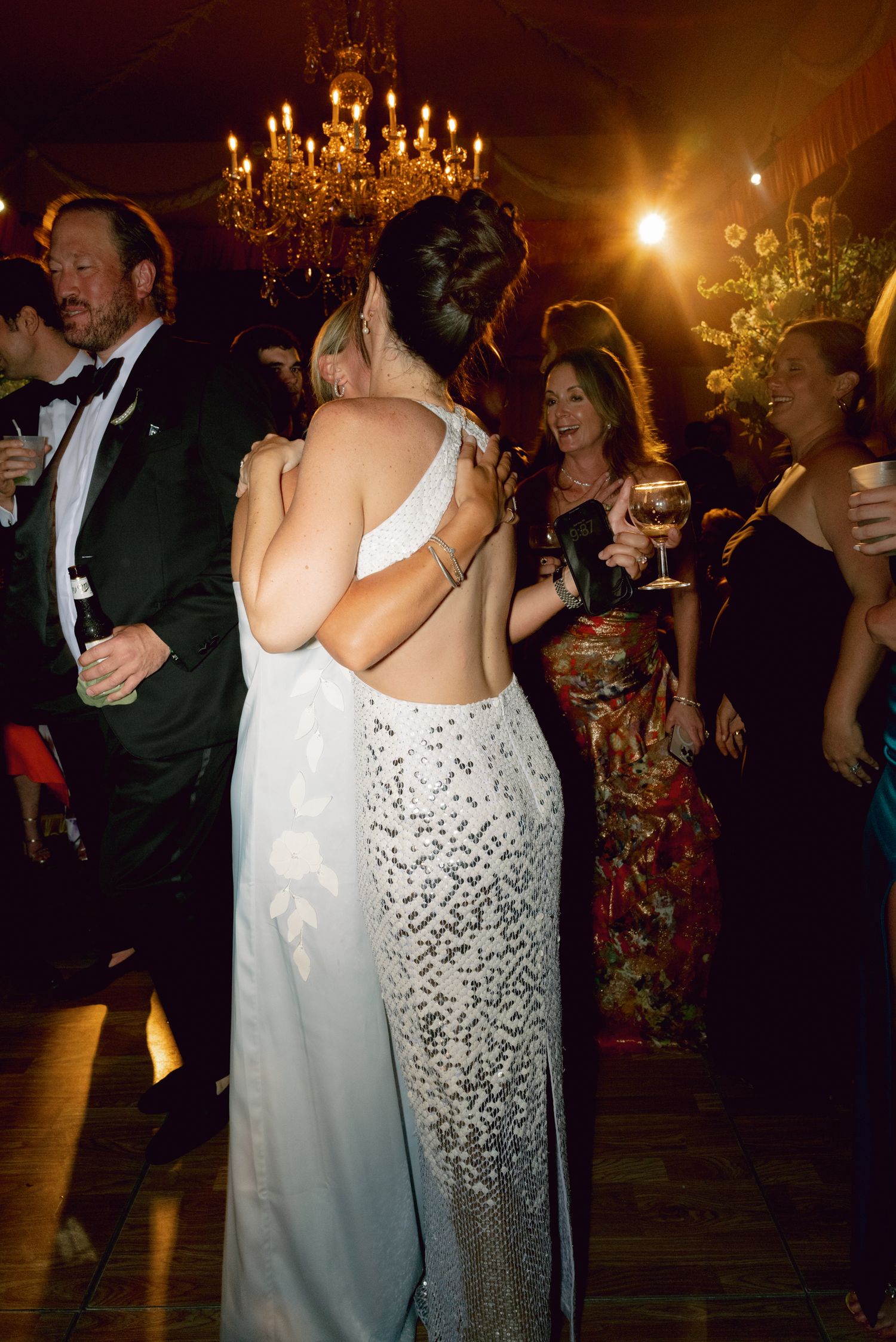 Two women embrace on a wedding dance floor under a chandelier, one wearing a sequined backless gown.
