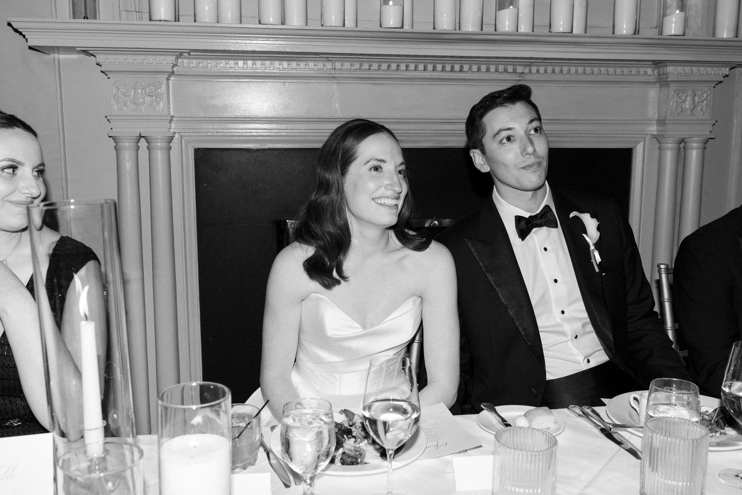Bride and groom smiling at sweetheart table during wedding reception in black and white photo.