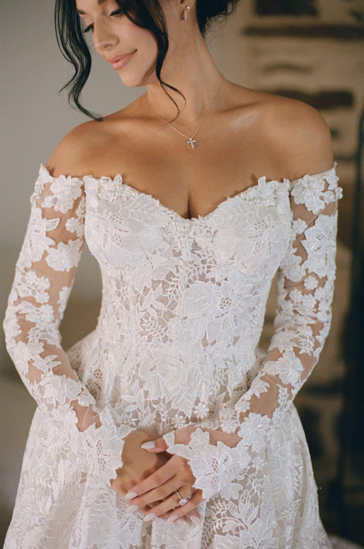 Bride in off-shoulder long sleeve lace wedding gown with floral appliques and cross necklace.