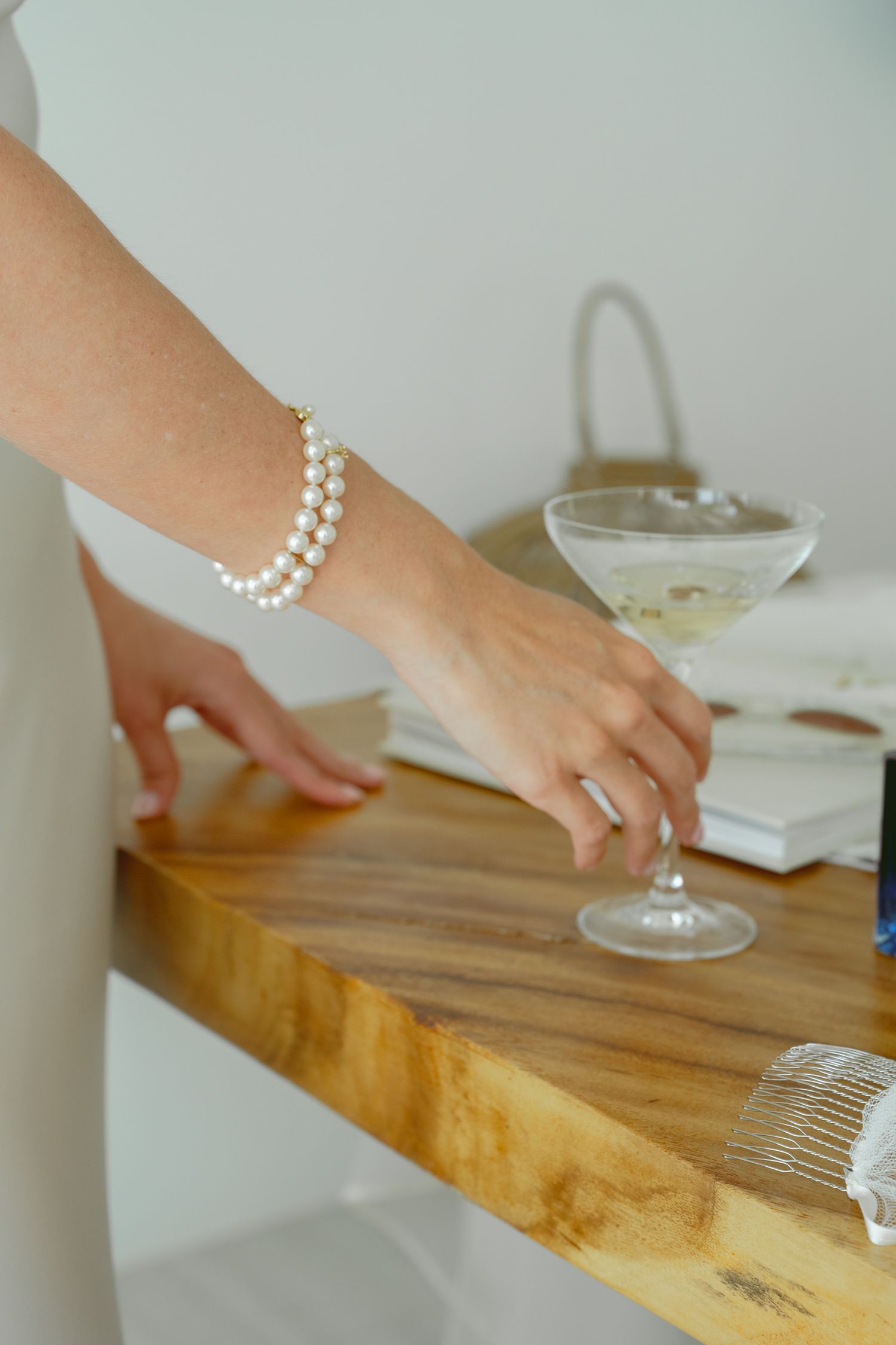 A hand wearing a pearl bracelet pours salt into a small glass bowl on a wooden cutting board.
