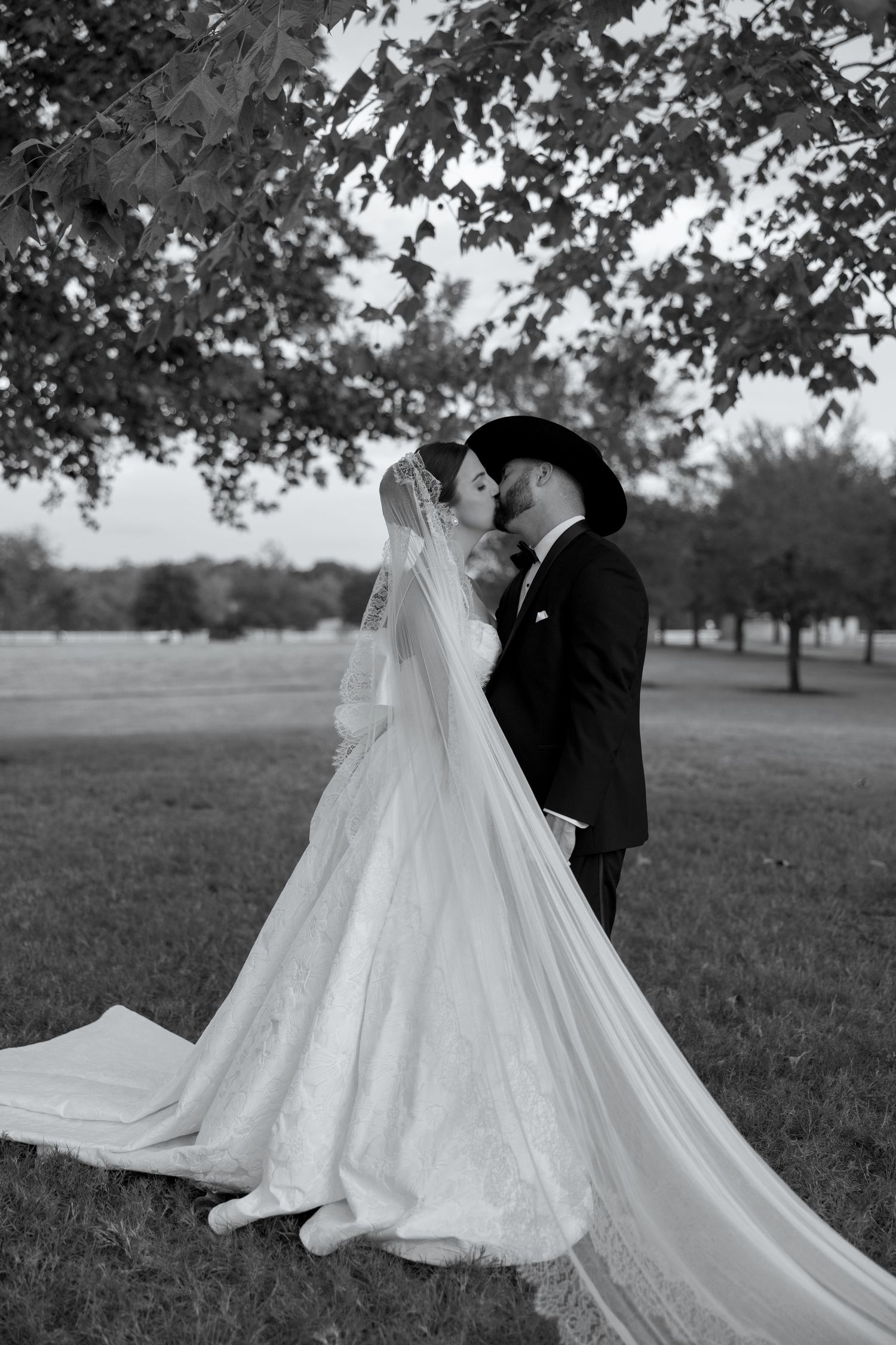 A black and white wedding photograph shows a couple in formal attire standing together in a scenic outdoor field.