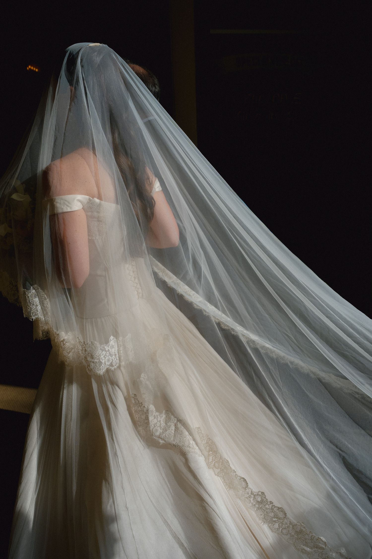 Bride in an elegant off-shoulder wedding gown with lace trim, draped in a flowing cathedral veil against dark background.