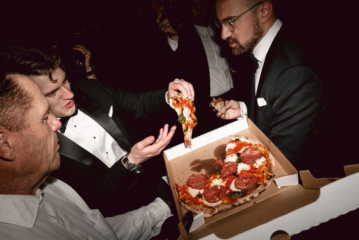 Men in tuxedos sharing a pepperoni pizza from a cardboard box at a formal evening event.