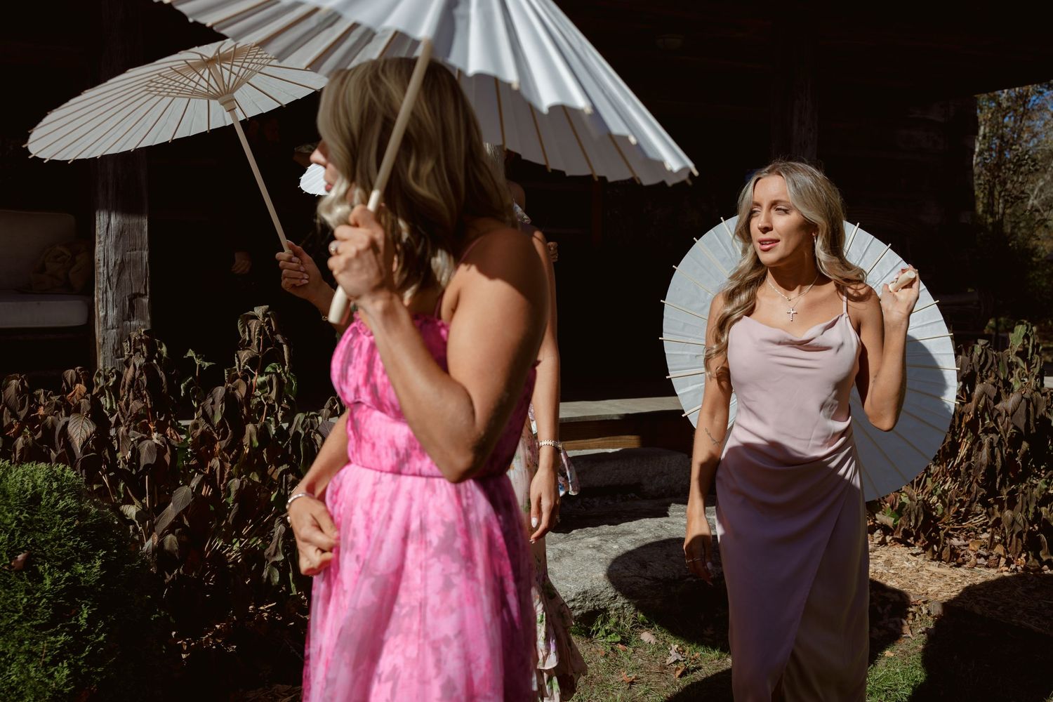Two women outdoors, one in pink floral dress holding white parasol, another in lavender outfit near decorative mirror.