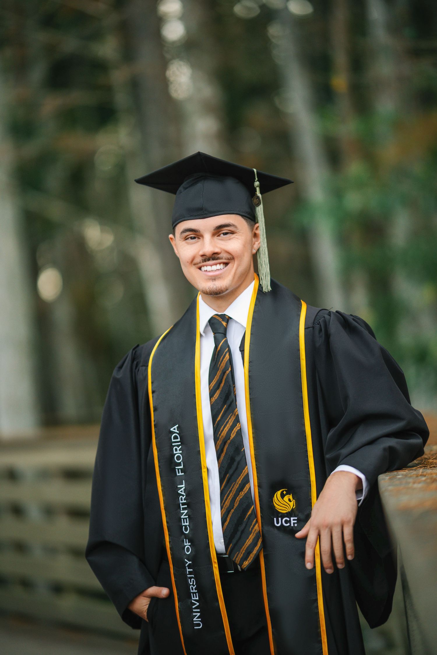 Smiling UCF graduate in black cap and gown with gold stole outdoors at University of Central Florida.