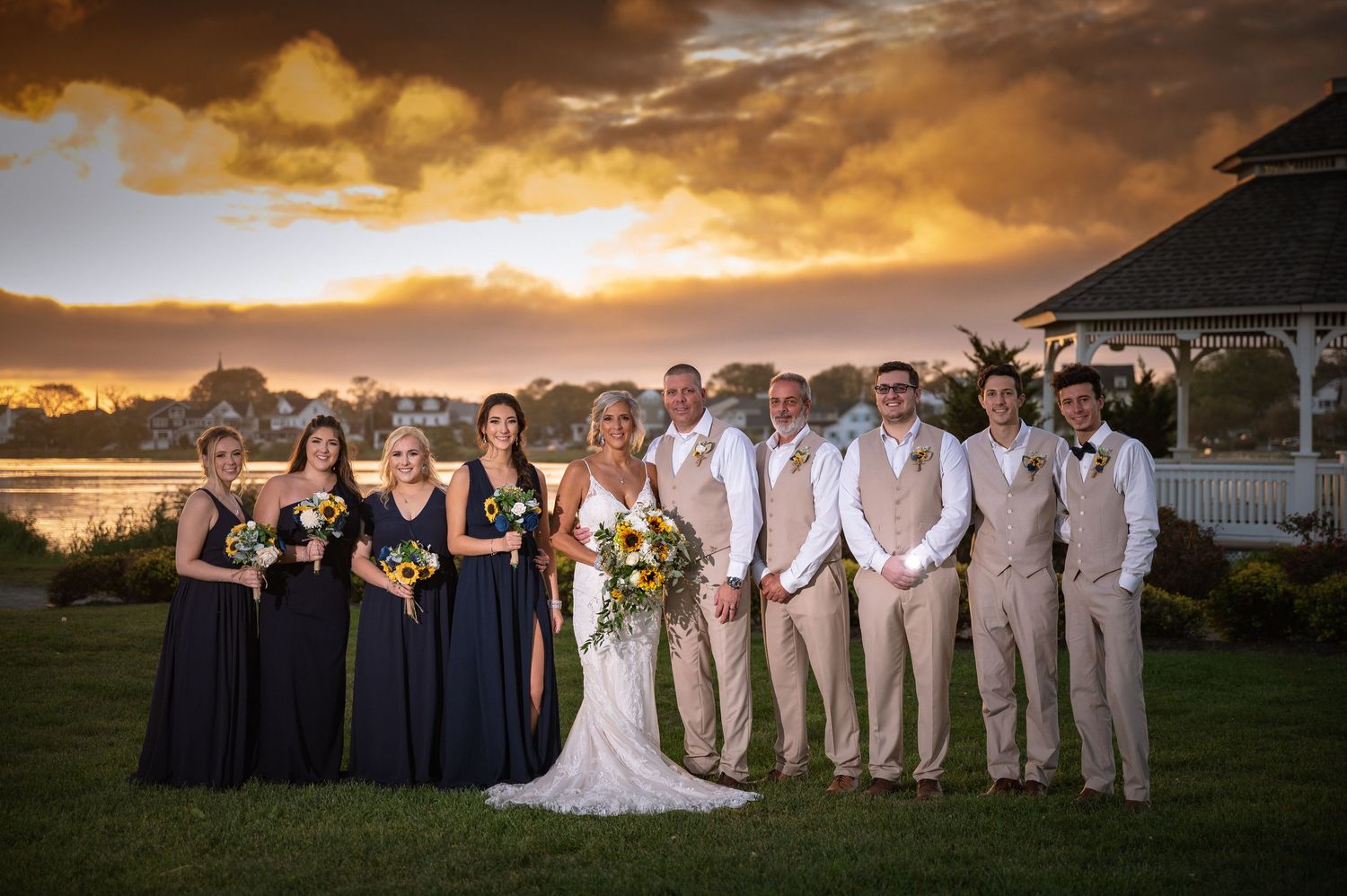 Wedding party poses at sunset near a gazebo and waterfront, bridesmaids in navy, groomsmen in tan vests.