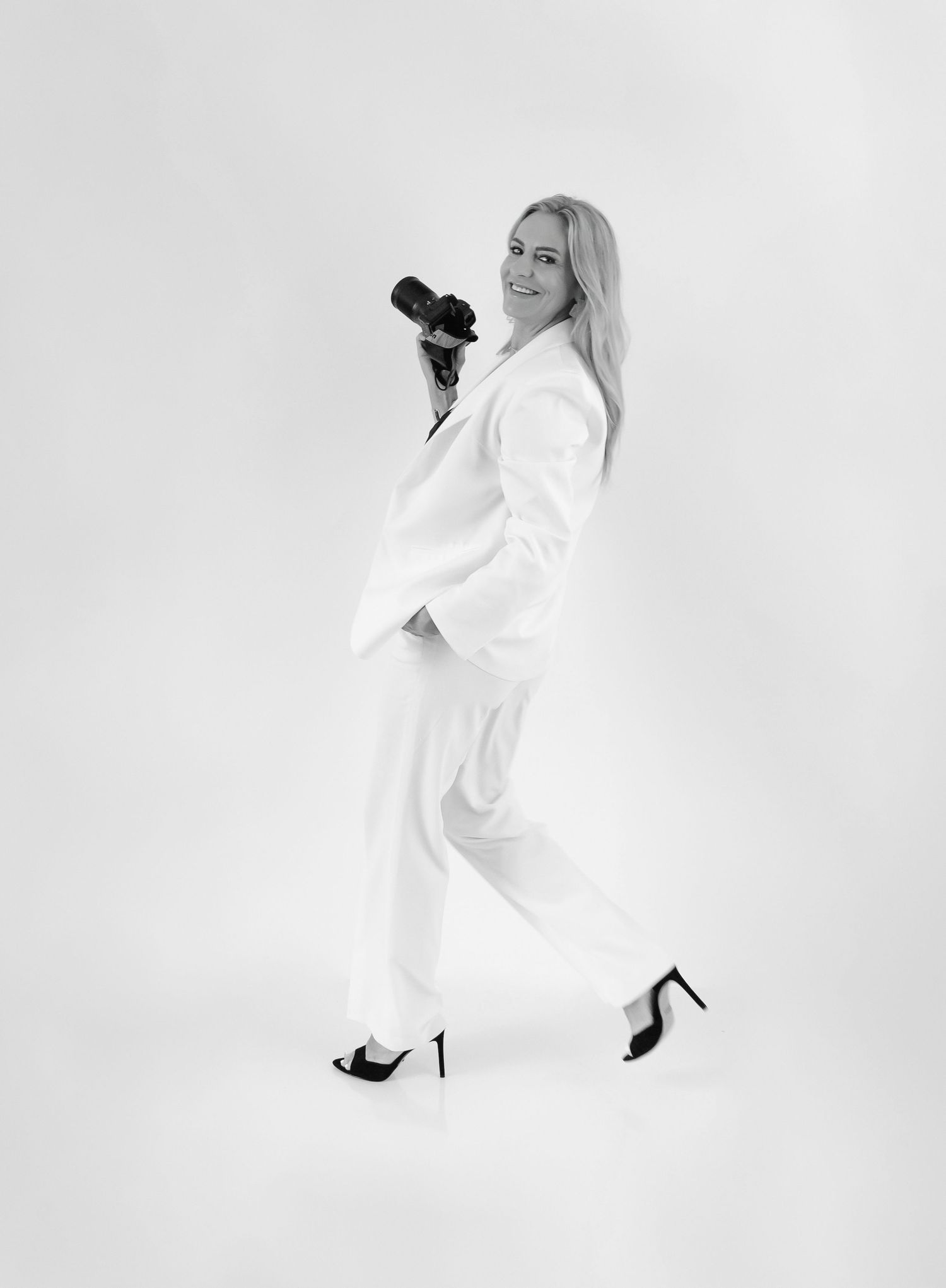 Woman in white suit holding camera, posing confidently in black heels, black and white studio portrait.