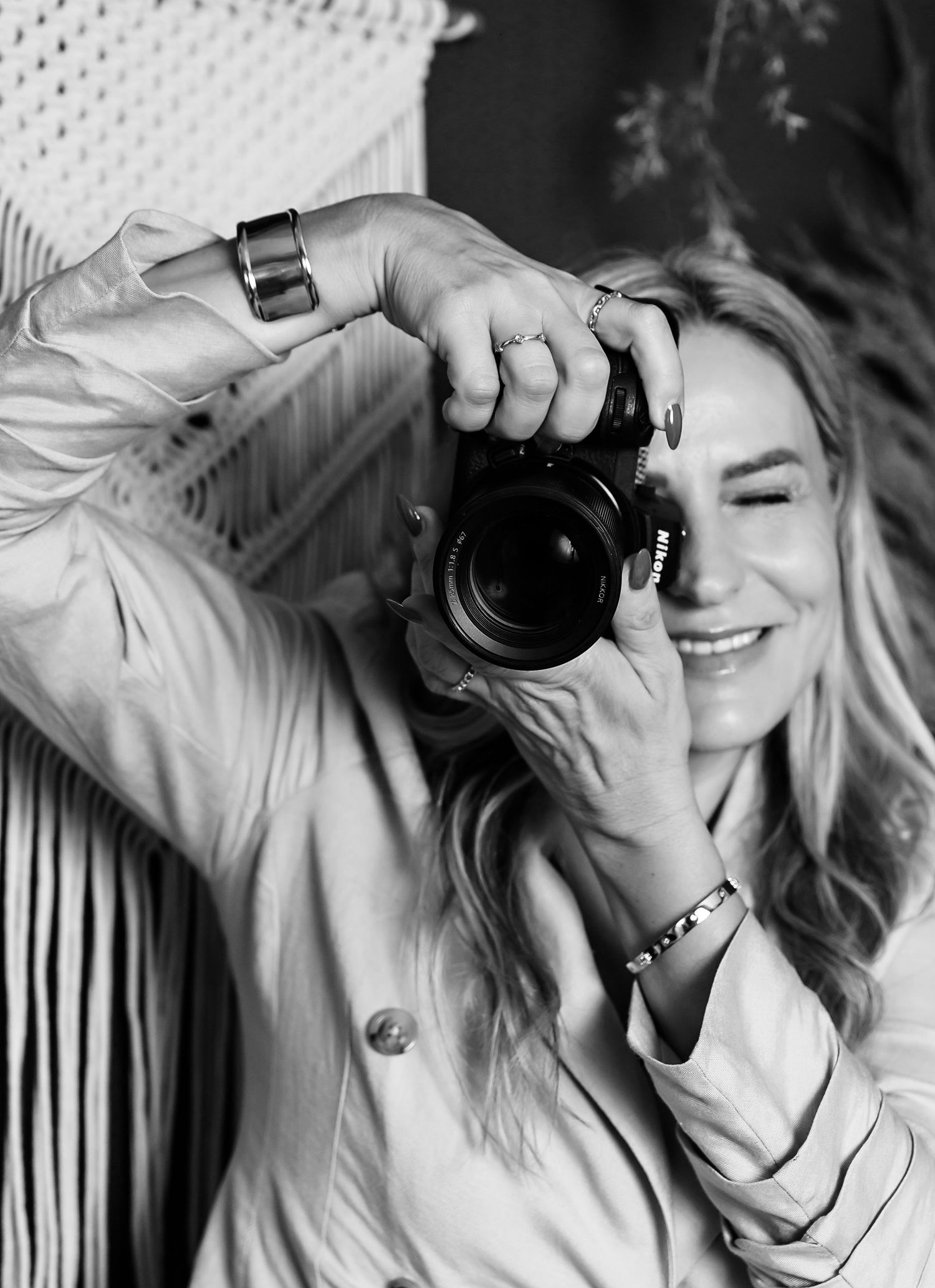 Black and white photo of a smiling woman holding a Nikon camera up to her eye, wearing rings and bracelets.