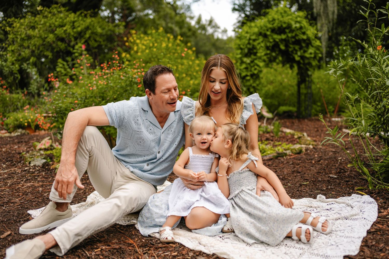 Family of four sitting on a blanket outdoors, parents smiling with two young daughters in a lush garden setting.