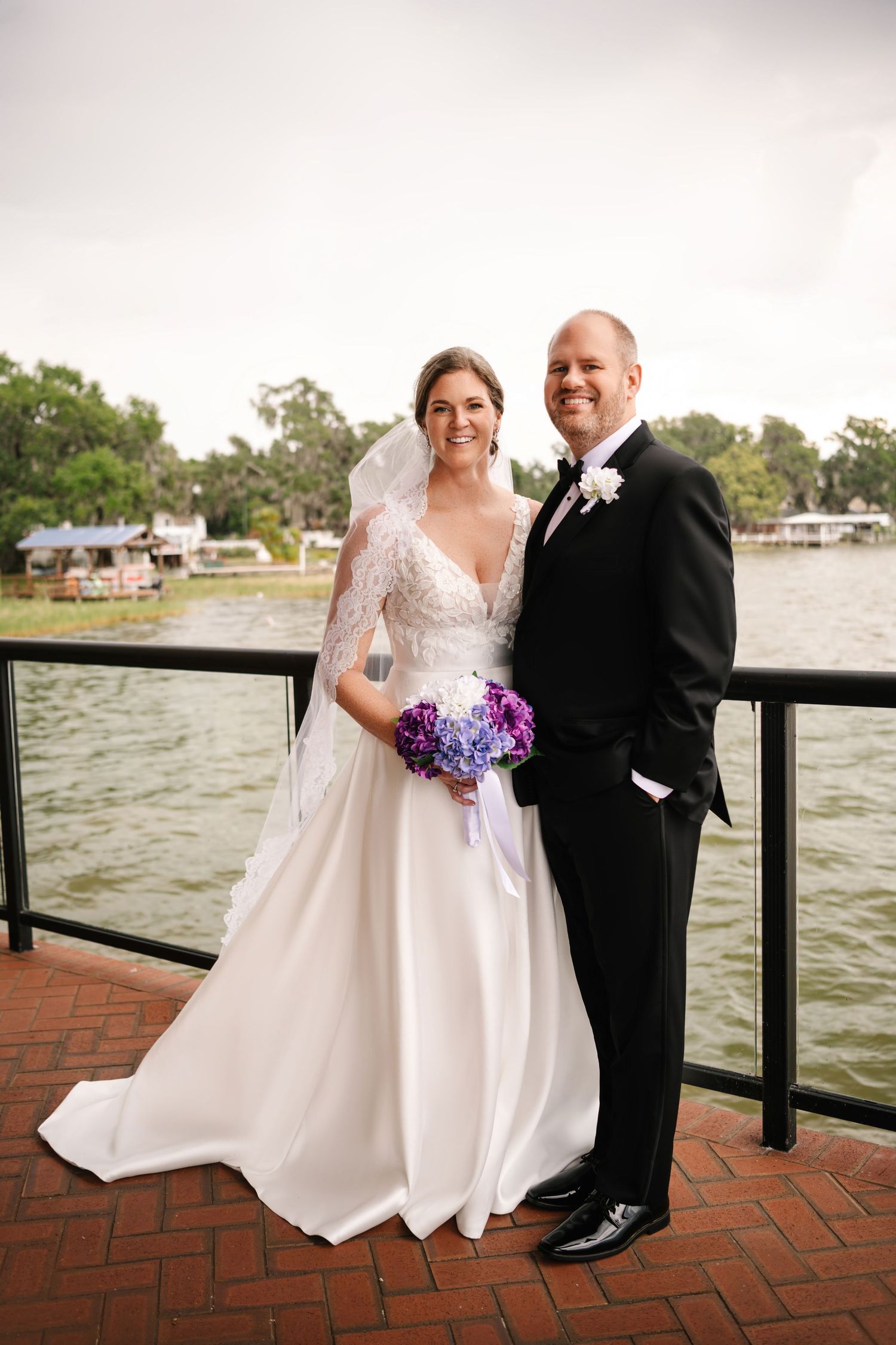 Bride and groom posing waterfront, she holds purple bouquet in lace gown with veil, he wears black tuxedo.