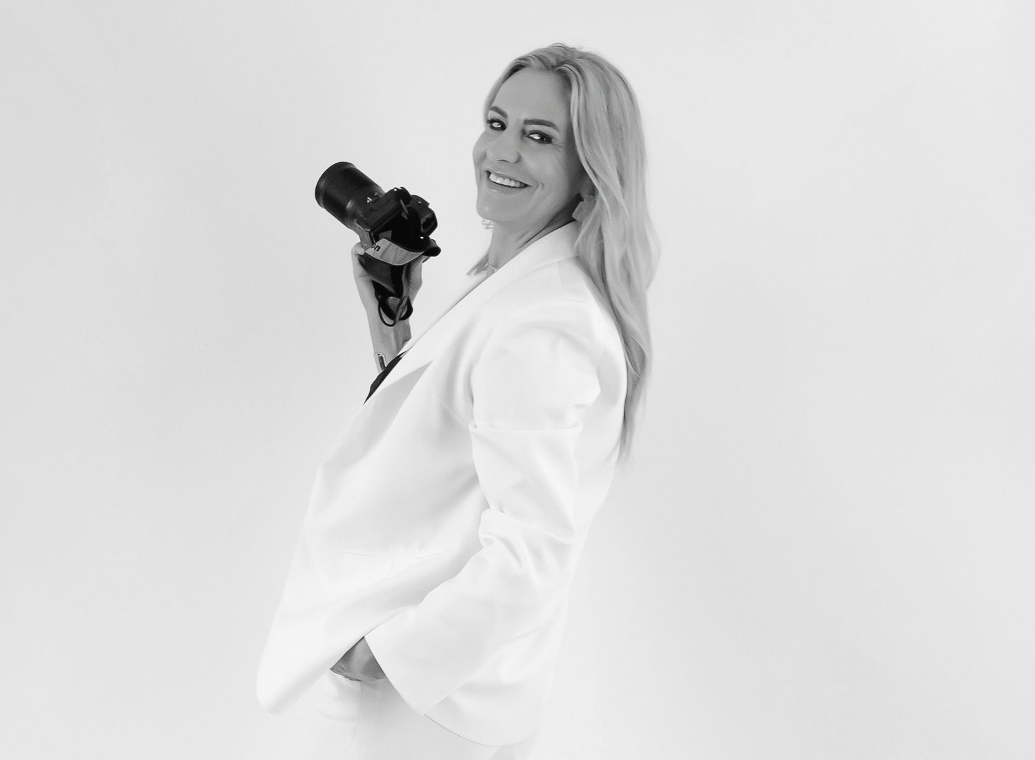Black and white photo of a smiling blonde woman in a white blazer holding a professional camera.