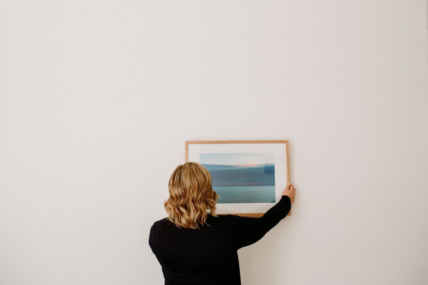 Someone in a black sweater hangs a framed coastal artwork on a white wall.