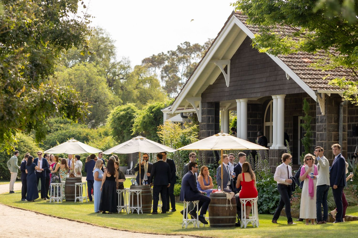 Wedding guests gather under white umbrellas and tents on a lush green lawn during an outdoor celebration.