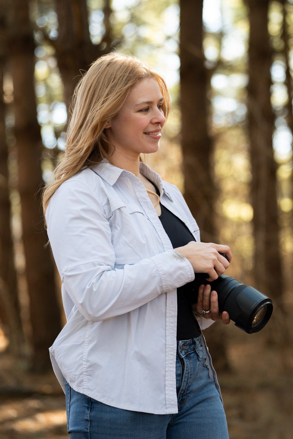 A person in a white button-down shirt and jeans stands in a sunlit forest setting.