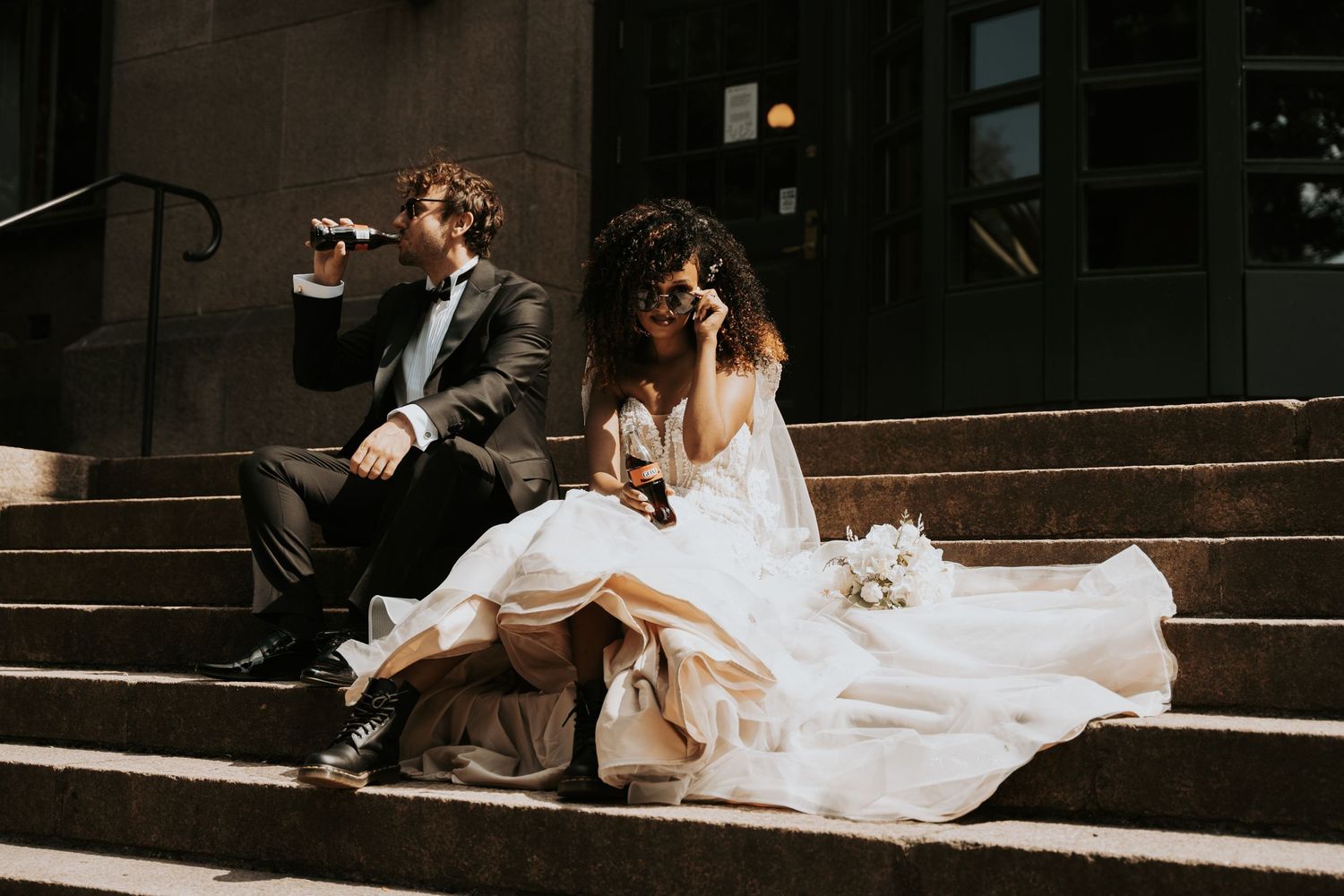 Newlywed couple sits casually on stone steps sharing drinks and laughing together in formal wedding attire.