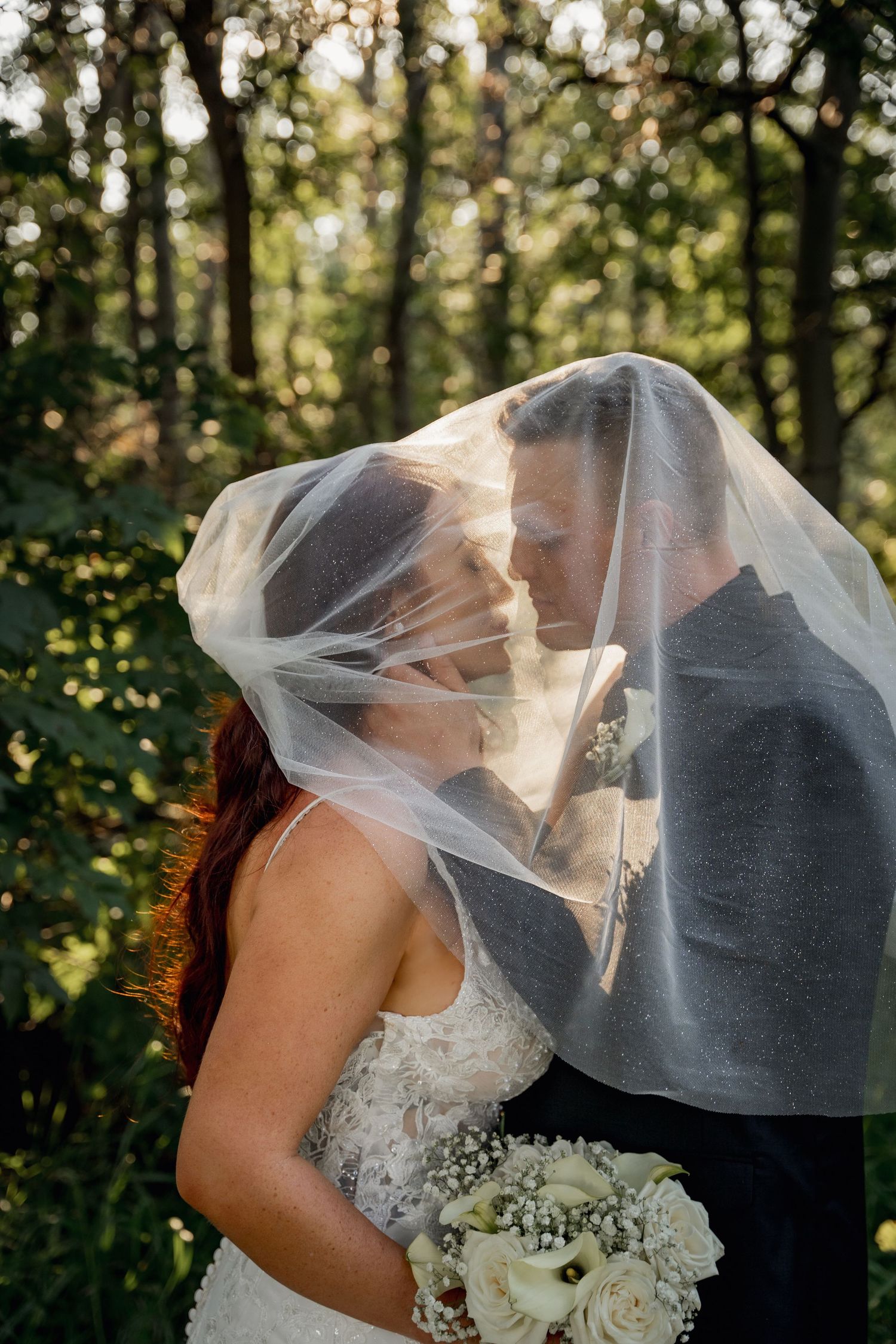 Romantic wedding moment captured under flowing veil in sunlit forest setting.