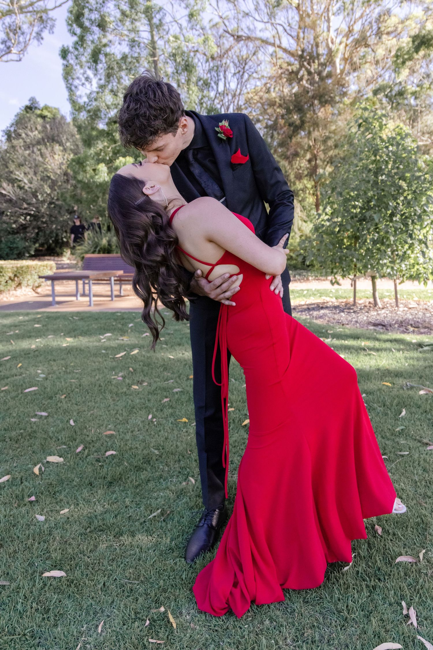 A couple in formal attire share a romantic dip pose on green grass, with the woman wearing a flowing red gown.