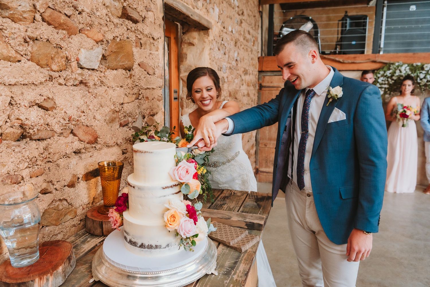 Newlyweds cut their elegant white wedding cake decorated with fresh flowers against a rustic stone wall.