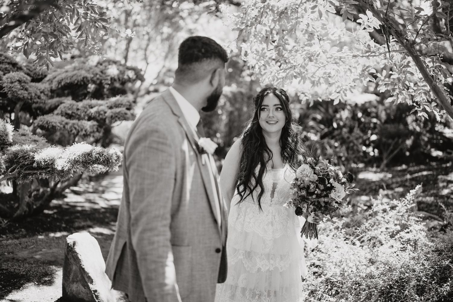 Black and white intimate moment at an outdoor wedding ceremony with trees and dappled sunlight creating a romantic atmosphere.