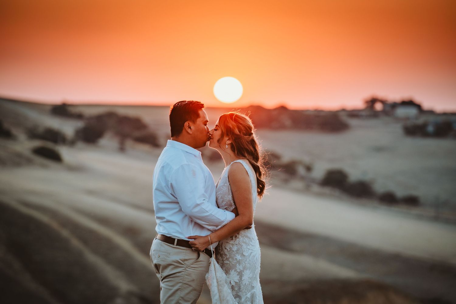 A couple shares a romantic moment at sunset on a hilltop with warm orange skies and rolling hills in the background.