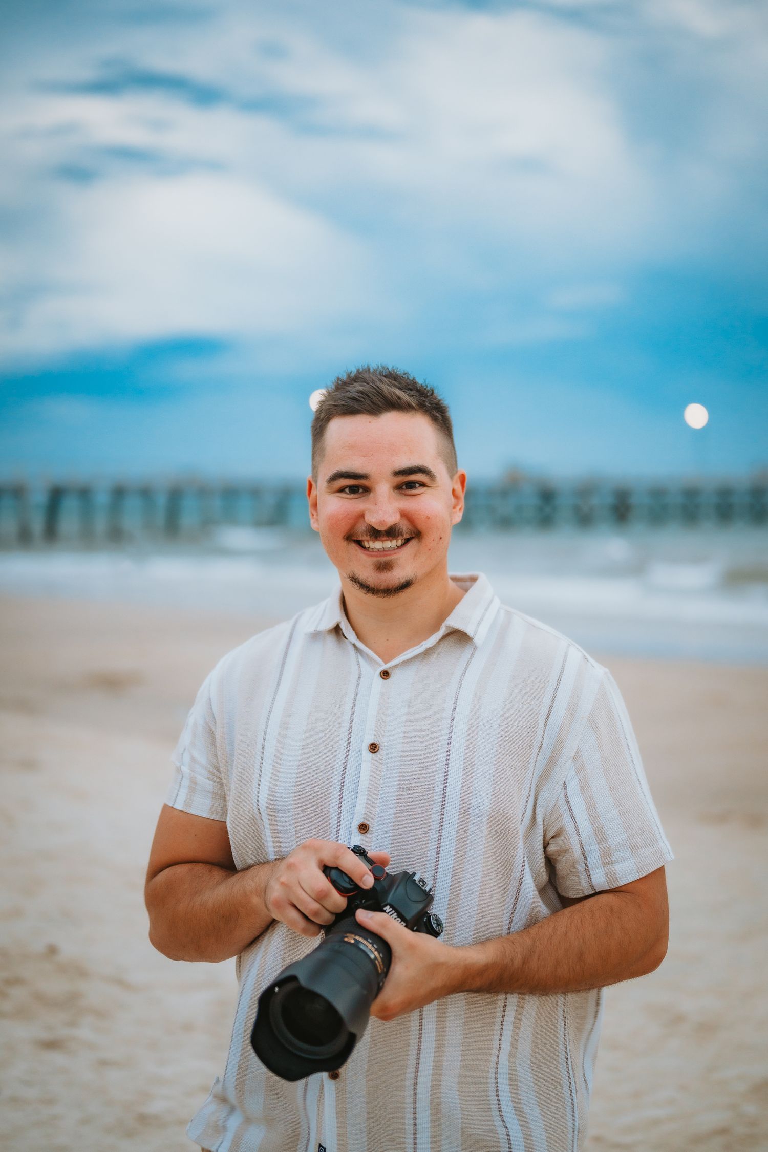 A photographer stands smiling on a beach holding a professional camera with a pier and ocean in the background.