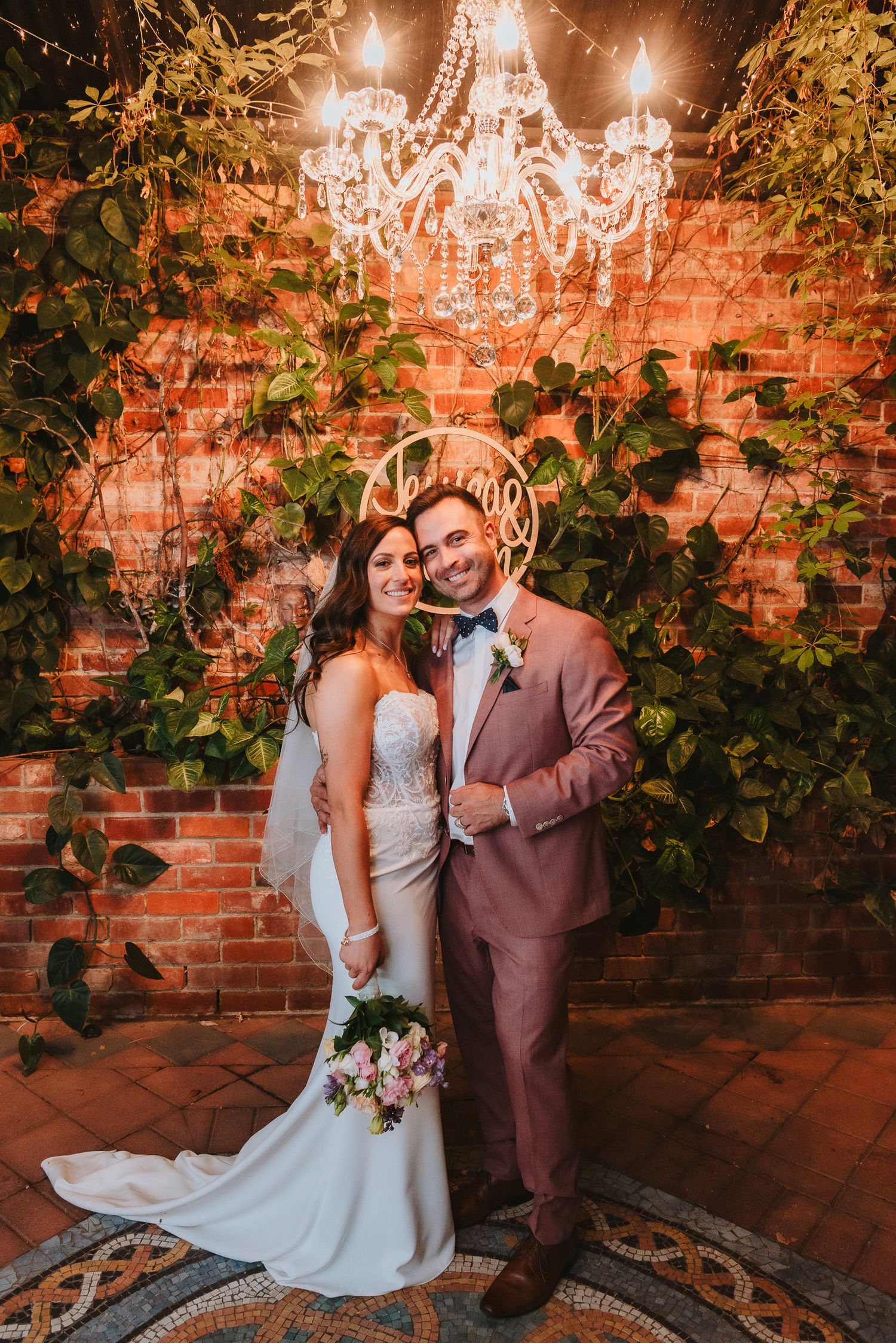 A romantic wedding portrait with a crystal chandelier, brick wall backdrop and greenery vines creating an elegant atmosphere.
