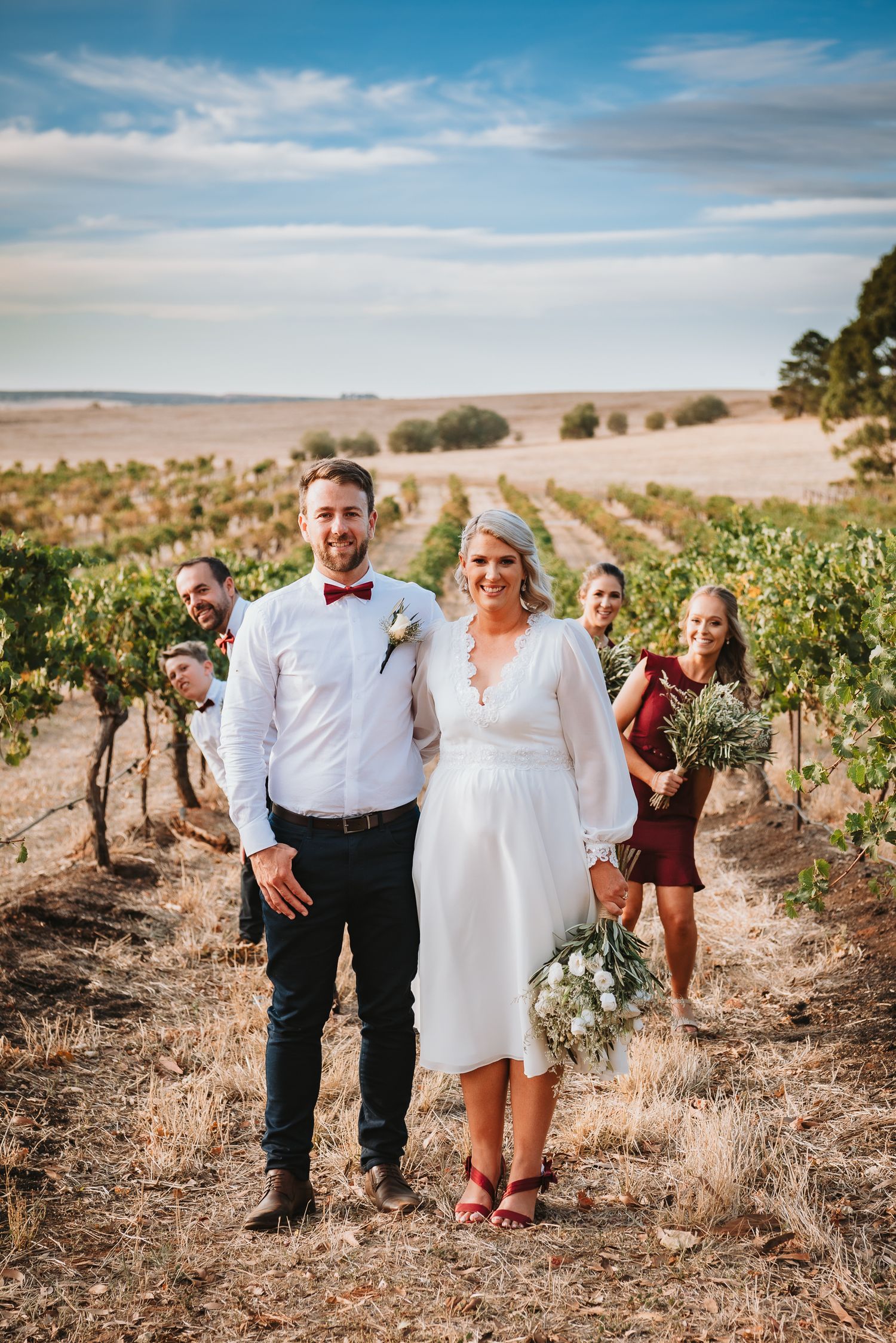 Wedding couple and guests stand in a vineyard on a sunny day with rolling hills and blue skies in the background.
