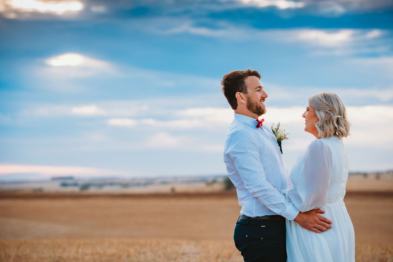 A joyful couple embraces and laughs together in a field under a bright blue cloudy sky.