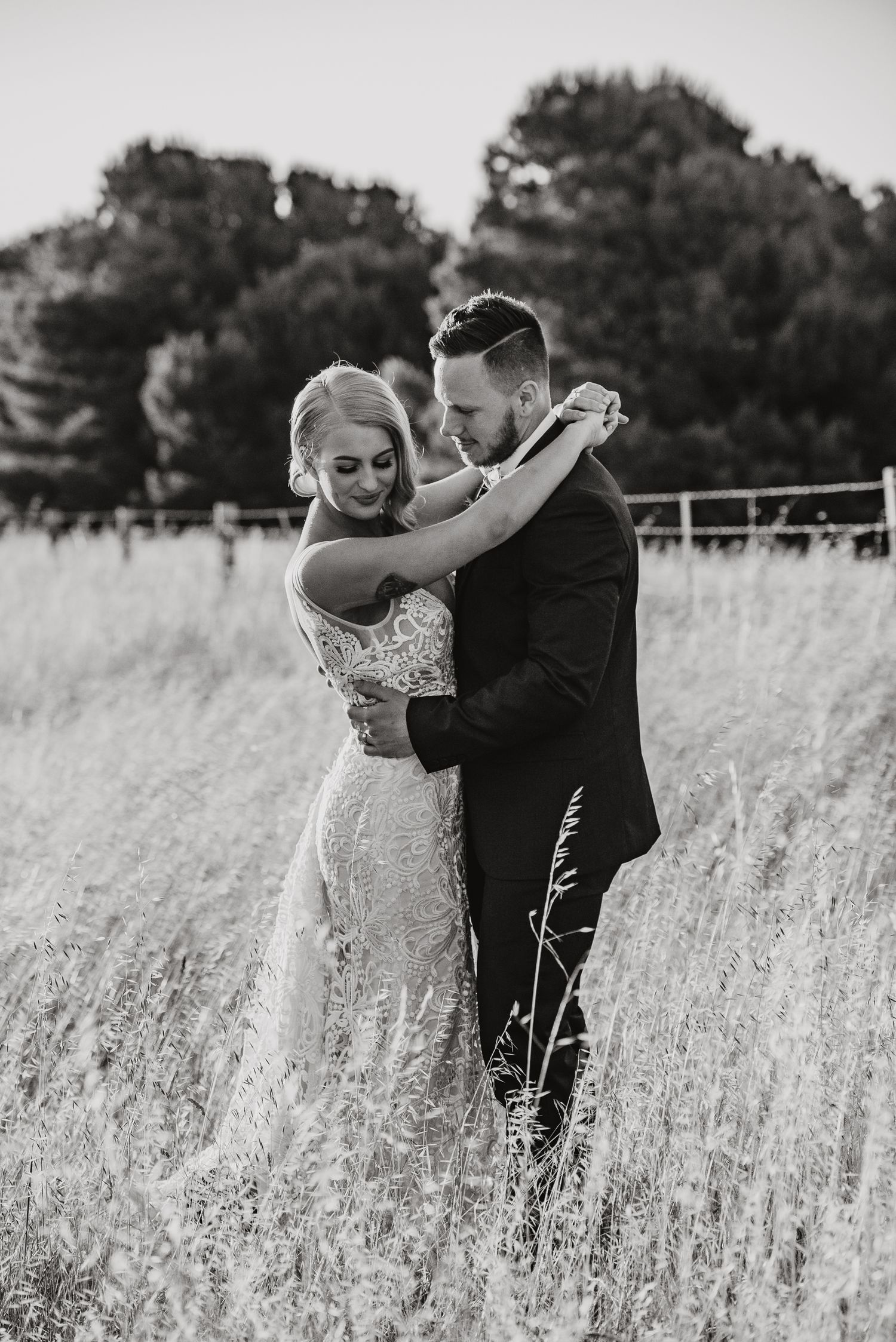 A romantic black and white wedding photo shows a couple embracing in a sun-lit meadow with tall grass and trees in background.
