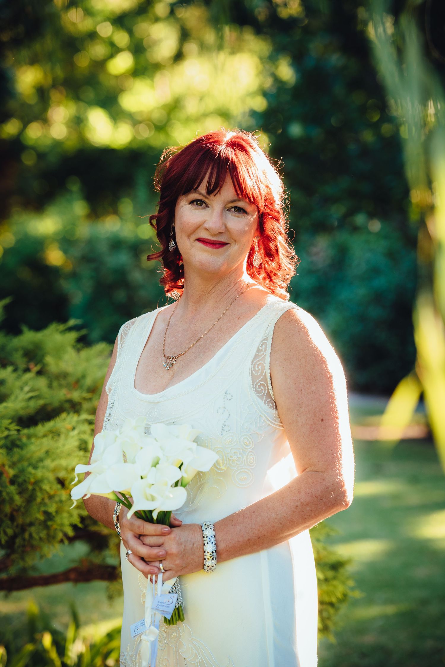 A bride in a white sleeveless dress holds a bouquet of white flowers in a sunlit garden setting.