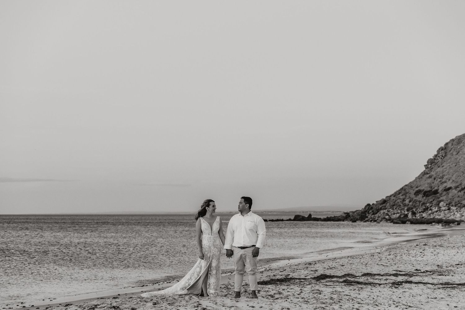 A romantic black and white beach scene with two people standing together by the shoreline and cliffs in the background.