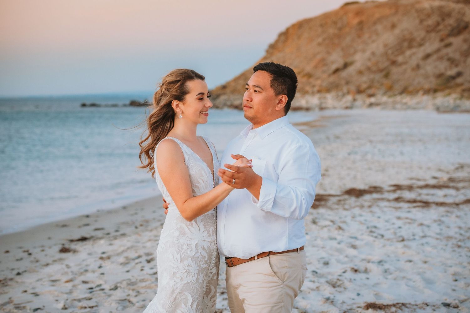 A couple in elegant white attire dances together on a scenic beach at sunset with cliffs in the background.