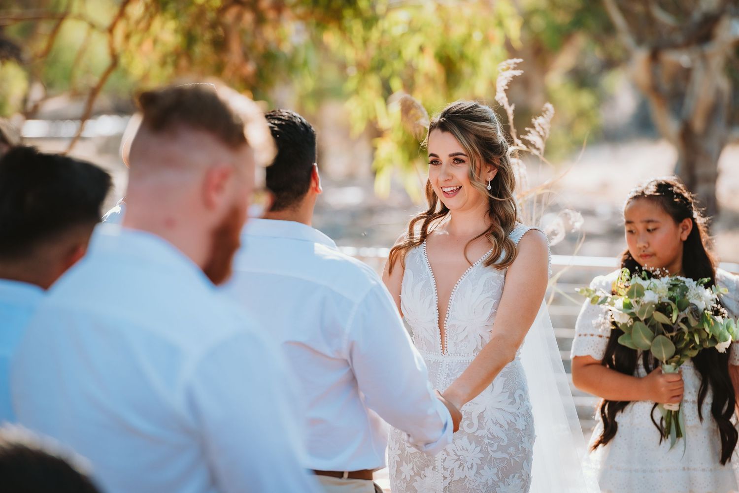 Wedding ceremony in progress outdoors with bride in white dress and wedding party under natural lighting.