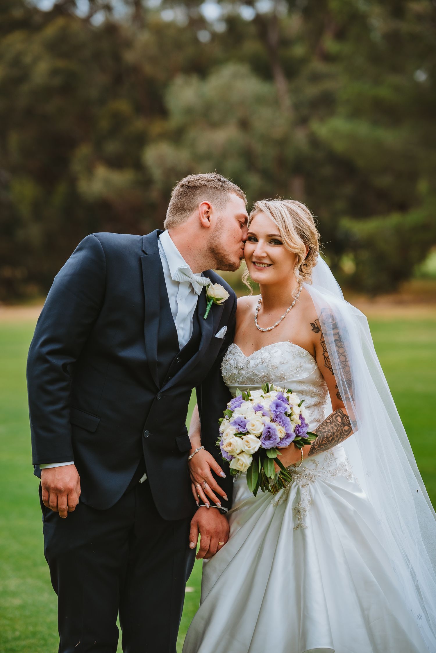 A romantic wedding moment captured outdoors with a beautiful white gown, purple bouquet and elegant navy suit.