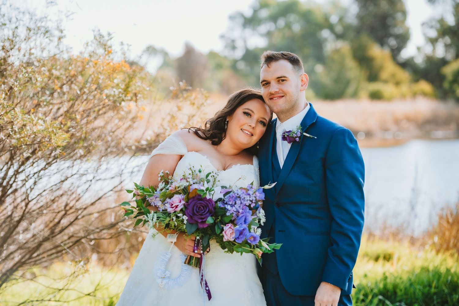 A couple in wedding attire embrace outdoors by a lake with the bride holding a purple and blue bouquet.