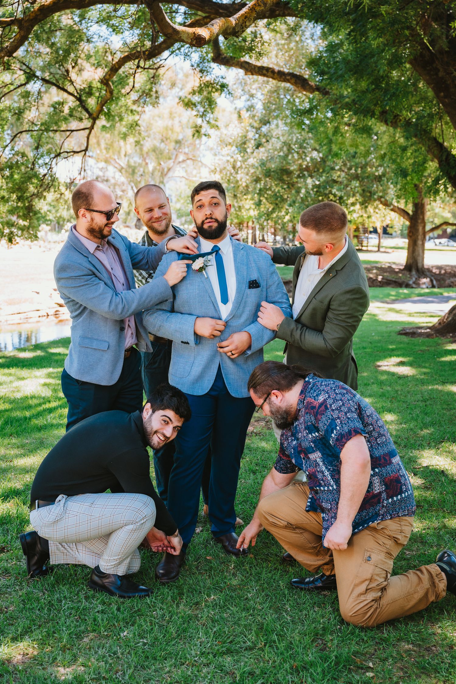 A groomsman in a blue suit playfully surrounded by his friends in a lush outdoor park setting.