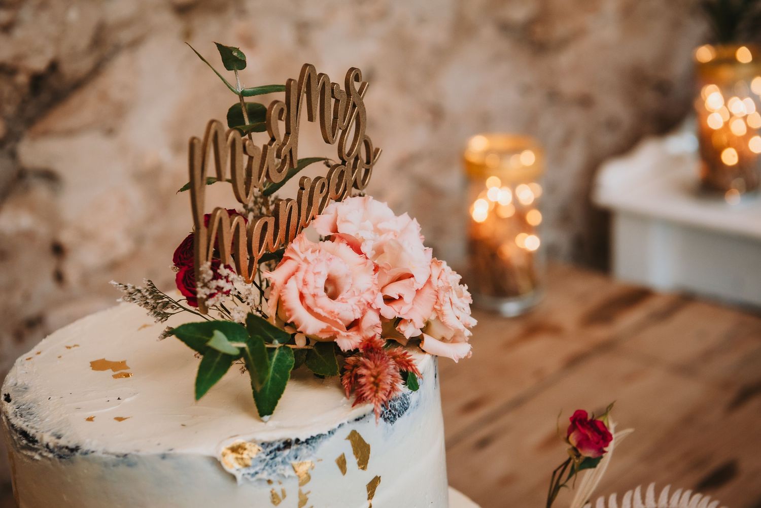 A white wedding cake decorated with pink roses and a wooden cake topper, illuminated by warm candlelight.