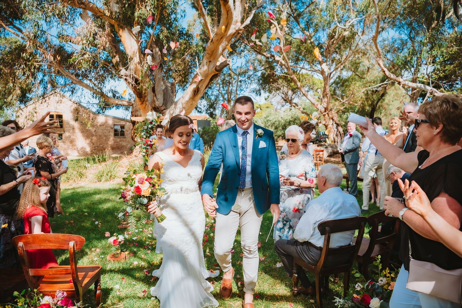 Wedding celebration takes place outdoors under eucalyptus trees with attendees throwing confetti on a sunny afternoon.